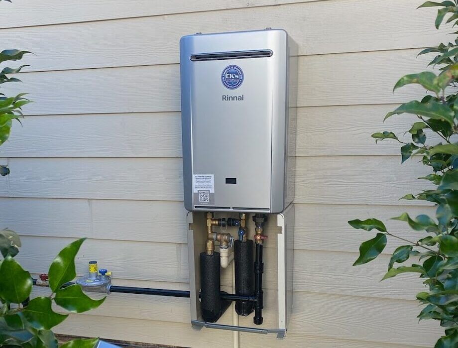 Outdoor wall-mounted tankless water heater on beige siding, with pipes and greenery nearby
