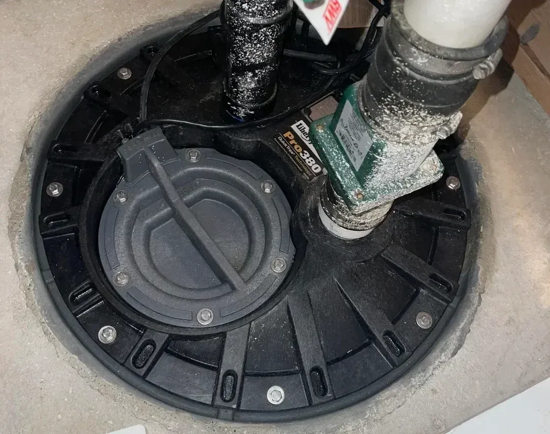 Black plastic sump pit with pipes and a round lid inside a concrete basin