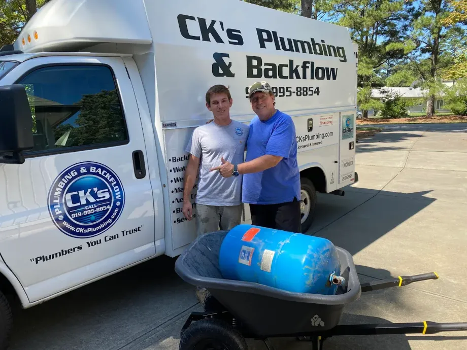Two men stand by a CK’s Plumbing & Backflow truck with a blue tank in a wheelbarrow on a sunny day