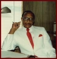 Man in white suit and red tie sitting at a desk, holding a pen, in an office setting.