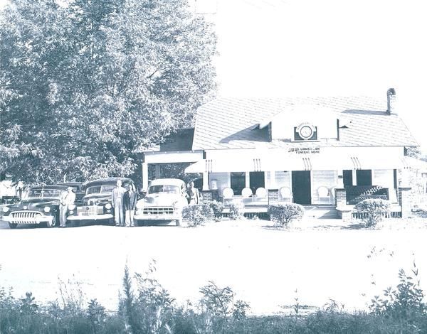 Vintage roadside house with parked cars and trees in front, seen in a washed-out black-and-white photo