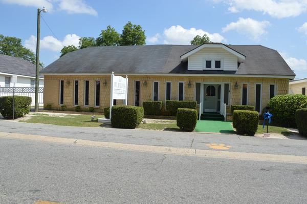 Small tan brick building with a dark roof, green ramp, and trimmed bushes along a street.