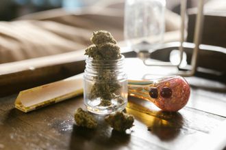 Marijuana buds in a glass jar, with a pipe and rolling paper on a wooden surface.