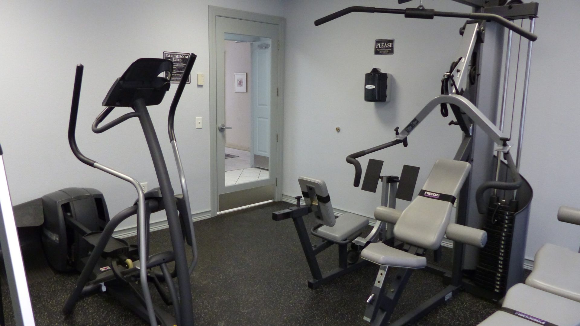 Small gym room with treadmill, weight machine, and exercise equipment against white walls.