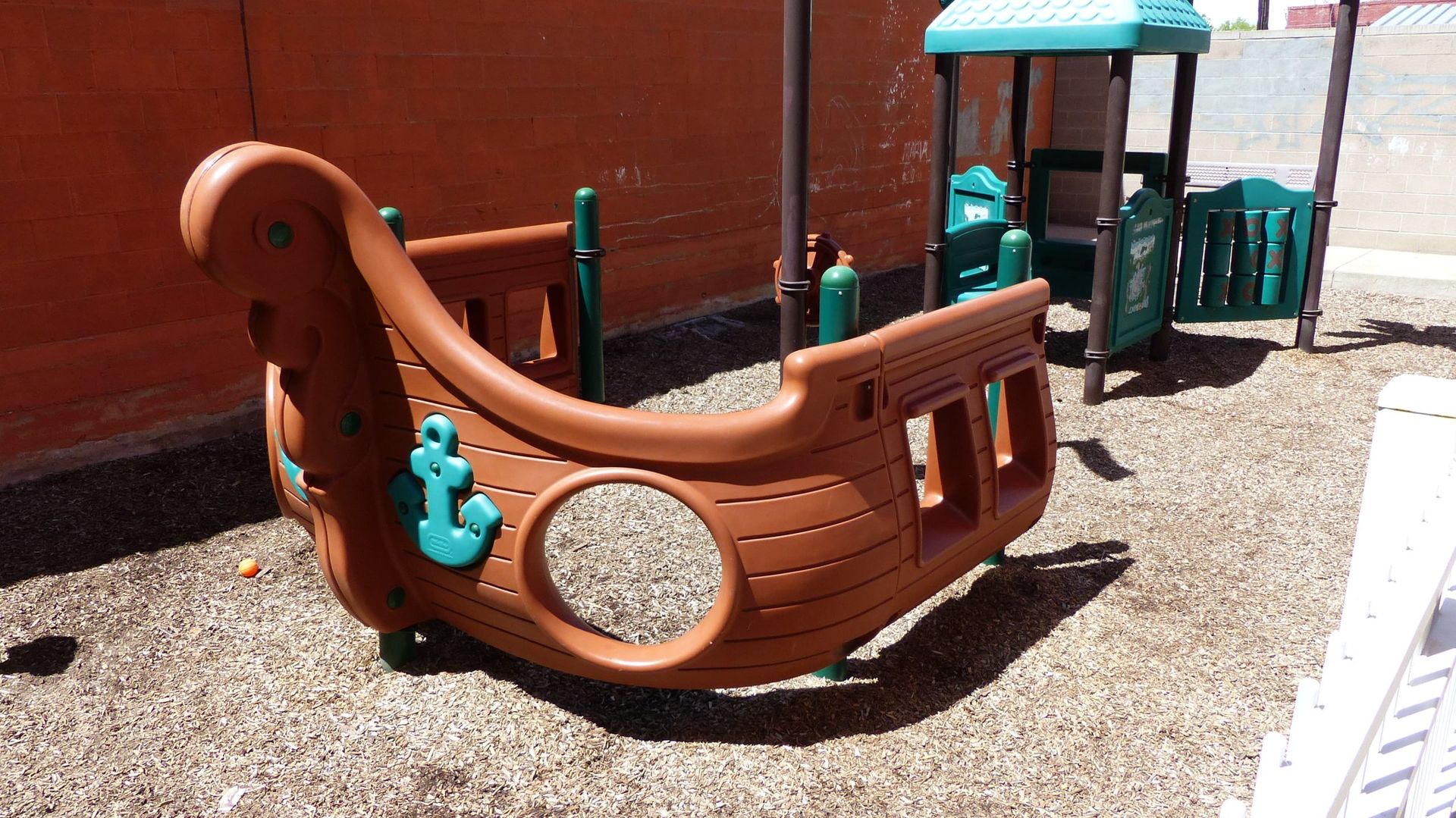 Brown pirate-ship playground climber with teal accents on a gravel play area beside a brick wall
