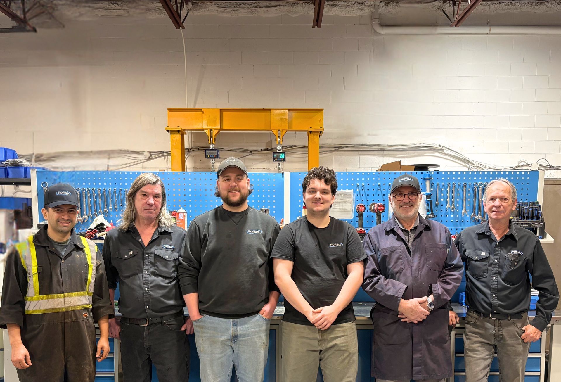 Six ouvriers, vêtus de leurs tenues de travail, se tiennent debout devant un panneau d'outils, à l'intérieur d'un atelier.