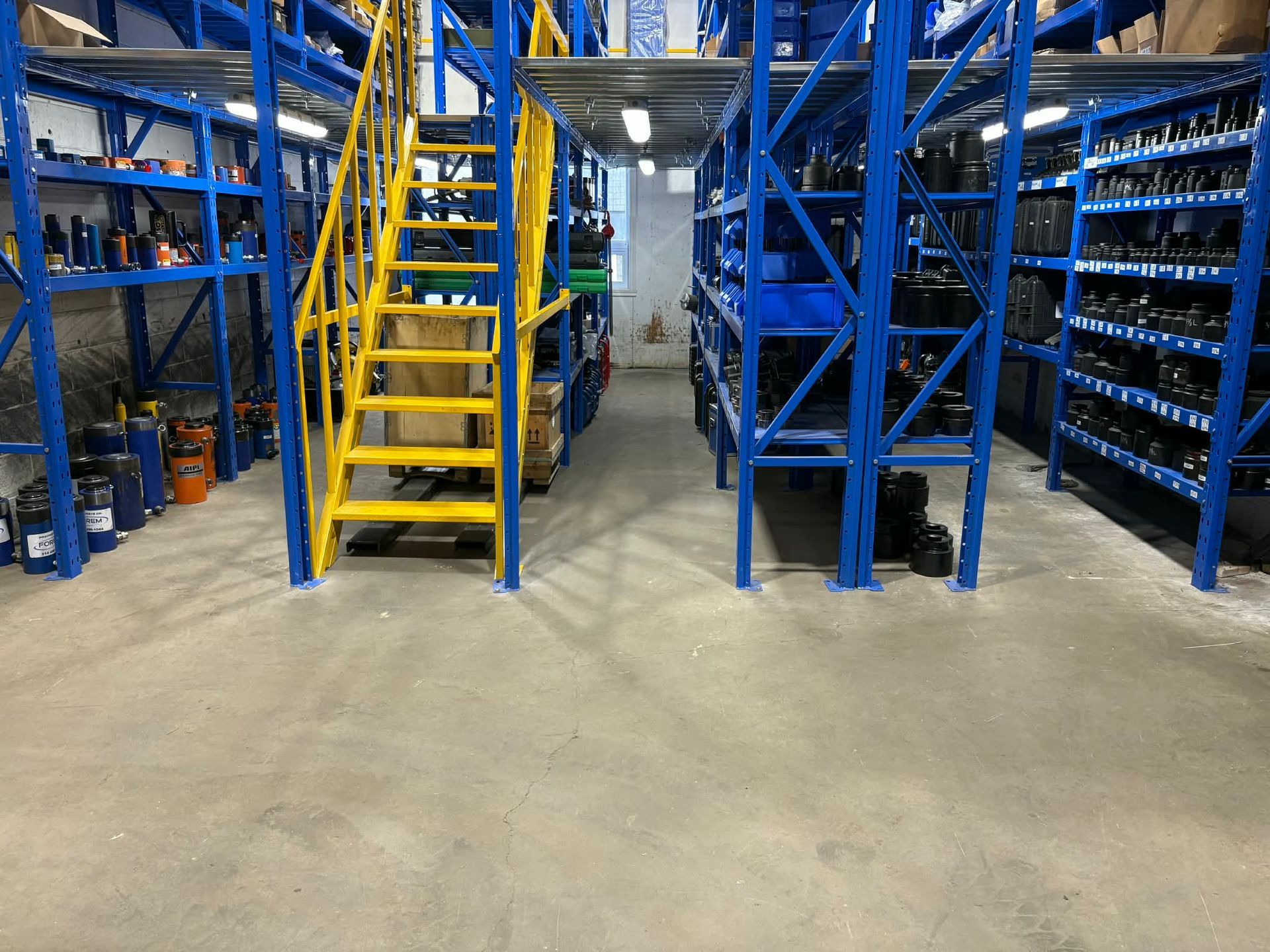 Warehouse storage area featuring blue metal shelving units, a yellow staircase, and a concrete floor.