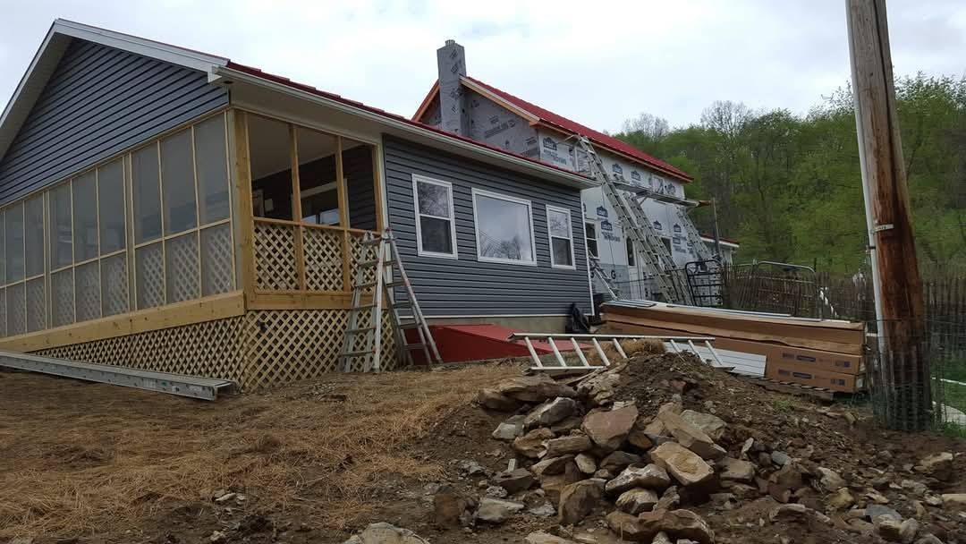 House under construction, gray siding, red roof, porch on the left, debris in foreground.