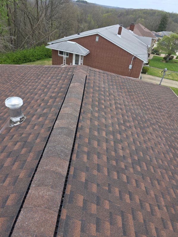 Rooftop view over a brown shingle roof toward a brick house and wooded hillside.
