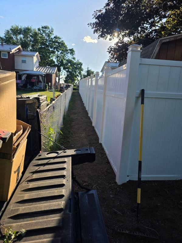 Narrow dirt path beside white vinyl fence with construction materials and a toolbox in a yard
