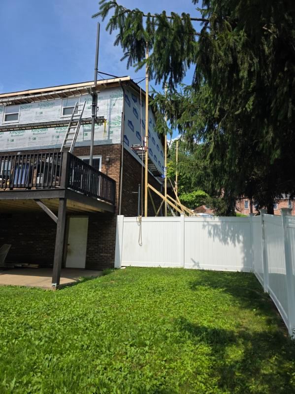 Backyard with green lawn, white fence, wooden deck, and tall trees beside a house under a blue sky
