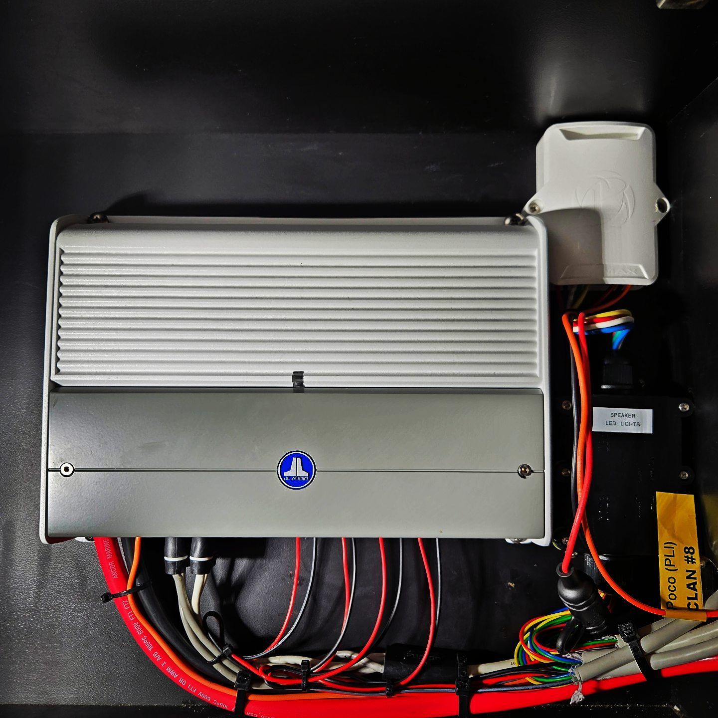 A silver JL Audio amplifier mounted in a black compartment with wiring and a white electrical component to the right.