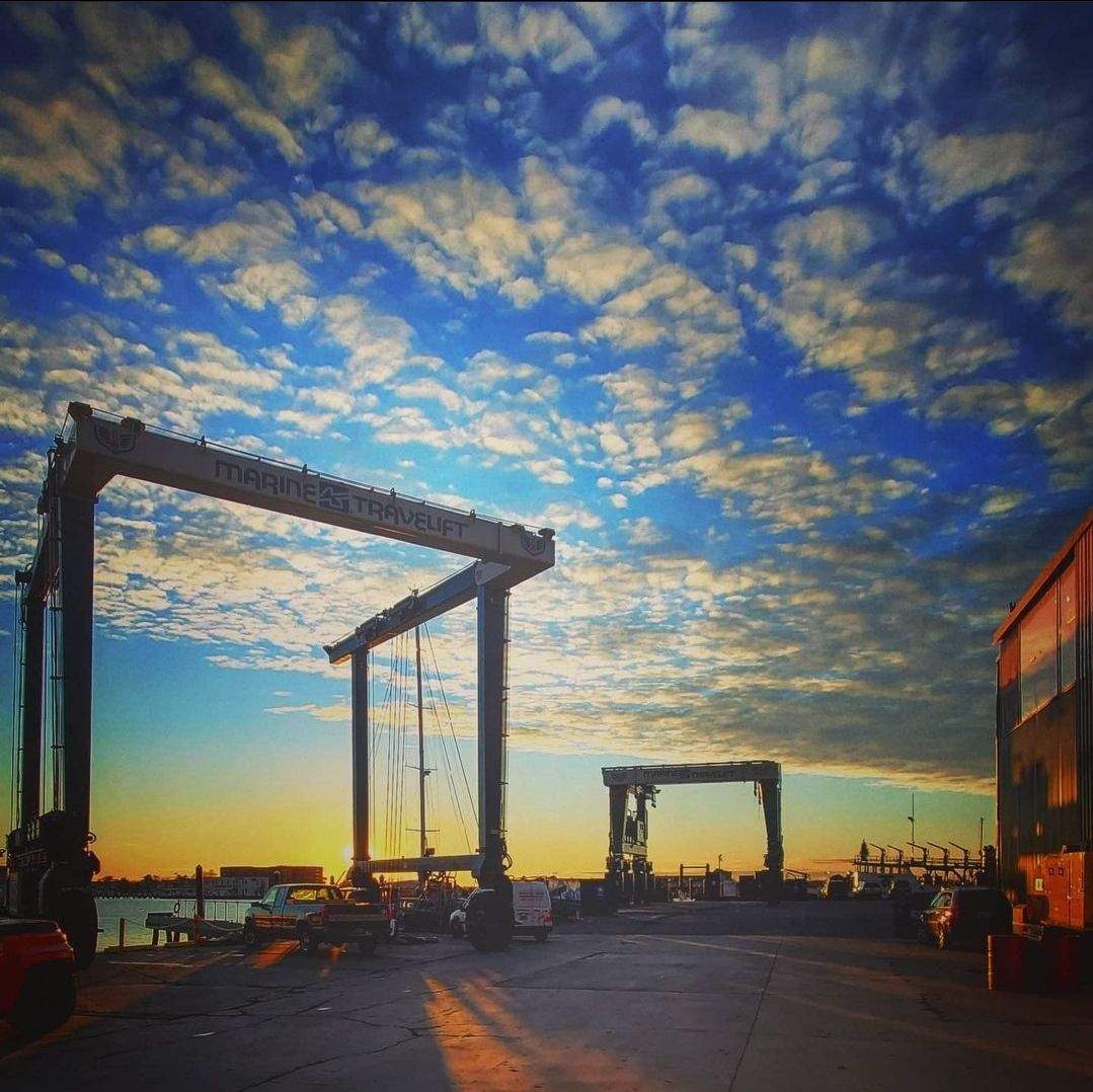 Two large boat-lifting cranes frame a sunset over a shipyard with a dramatic, textured sky.