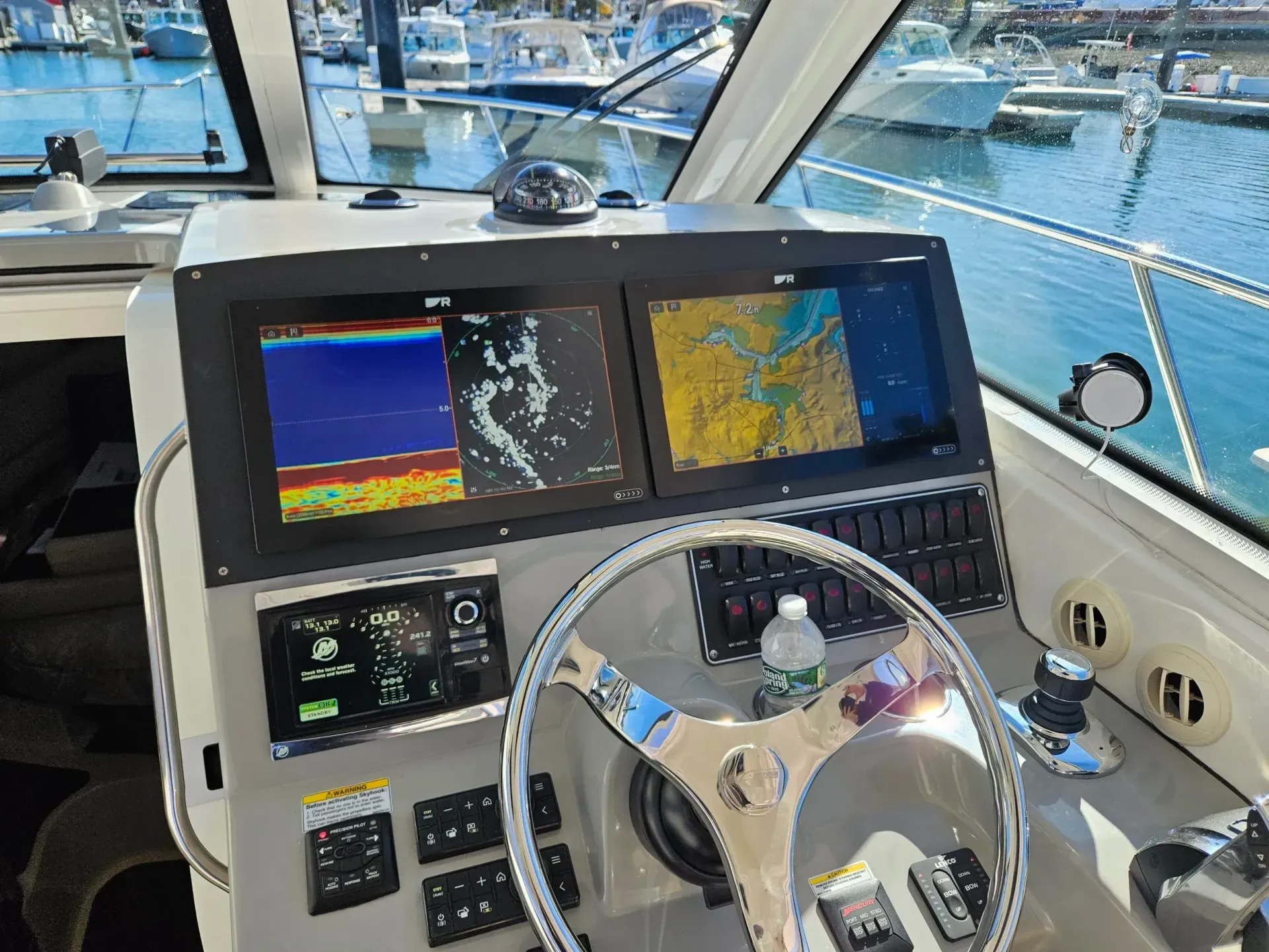 Boat helm station with dual marine navigation displays, a chrome steering wheel, and switches, overlooking a marina.