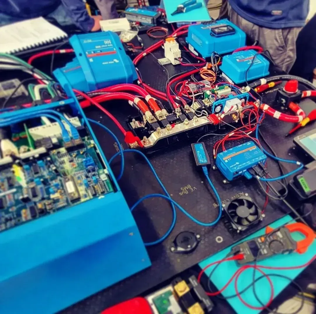 An electrical workbench with blue Victron Energy components, thick red cabling, busbars, a circuit board, and tools.