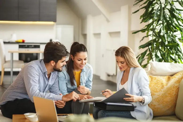 Three people discussing documents in a bright living room with a laptop and coffee table.