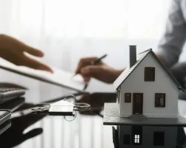 Hands exchanging a house key over a desk with a small model home, suggesting a property sale or rental deal