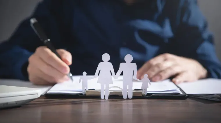 Hands writing in a notebook beside paper family figures on a desk, with a laptop in the background