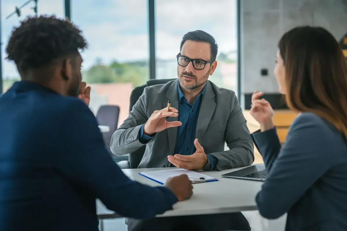 Three people in a meeting, discussing around a table in a bright office.