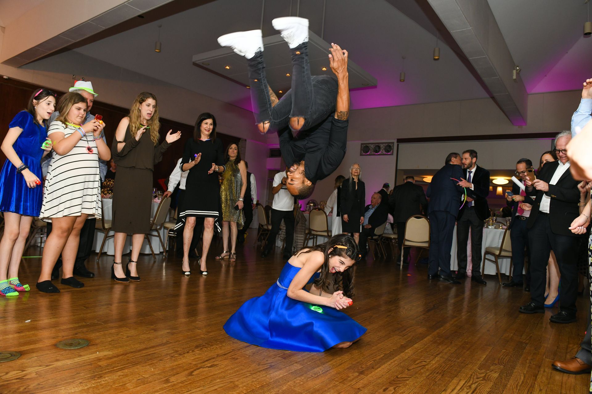 A person performs a backflip over a person kneeling in a blue dress at a party with guests watching.