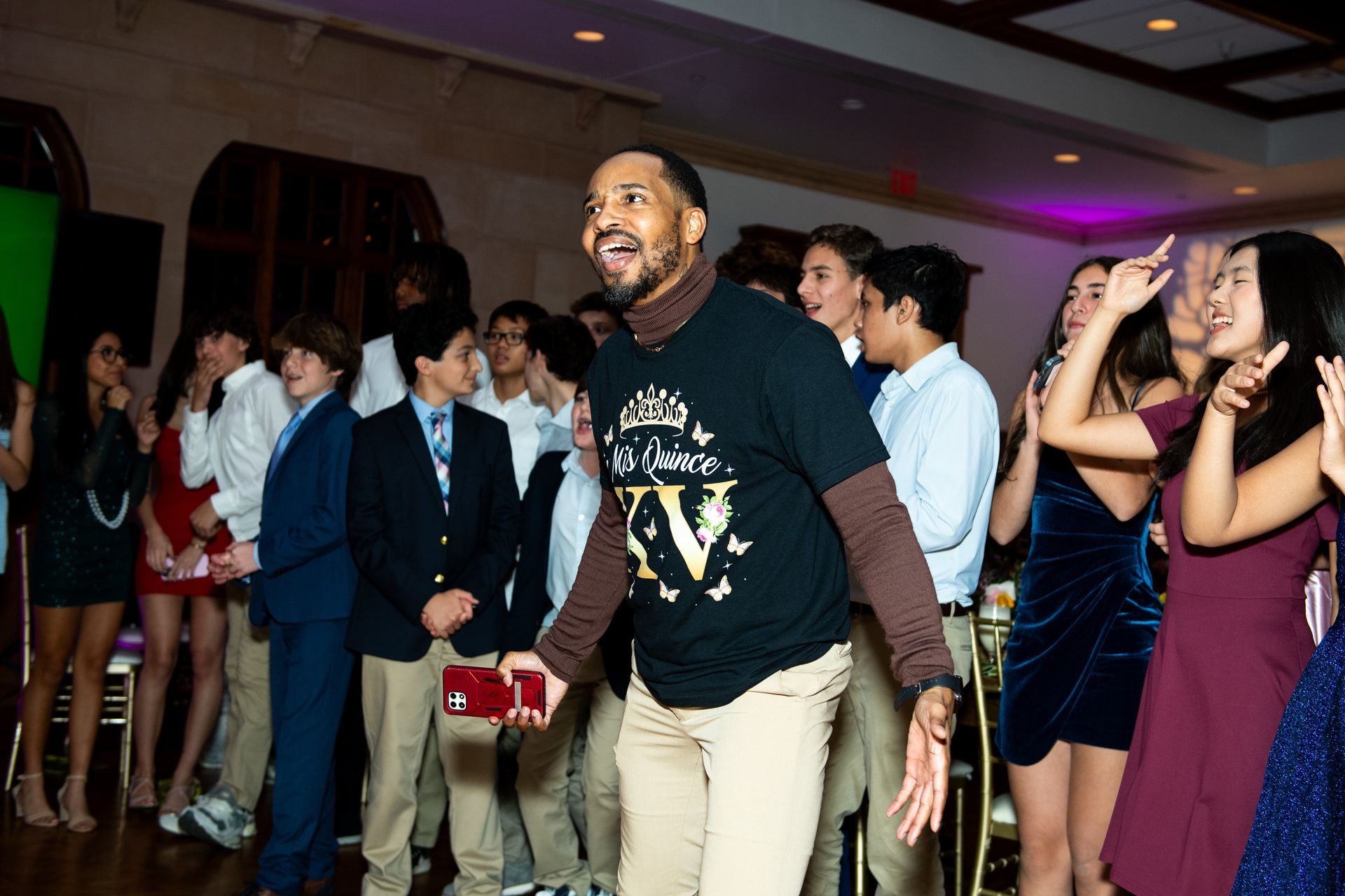 A man in a graphic shirt and khakis smiles excitedly at a party surrounded by people in semi-formal attire.