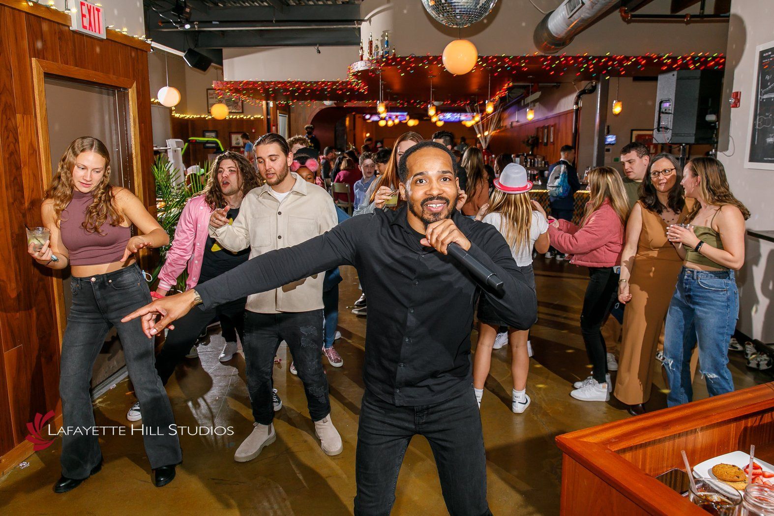 A person sings into a microphone at a crowded bar, with others dancing.