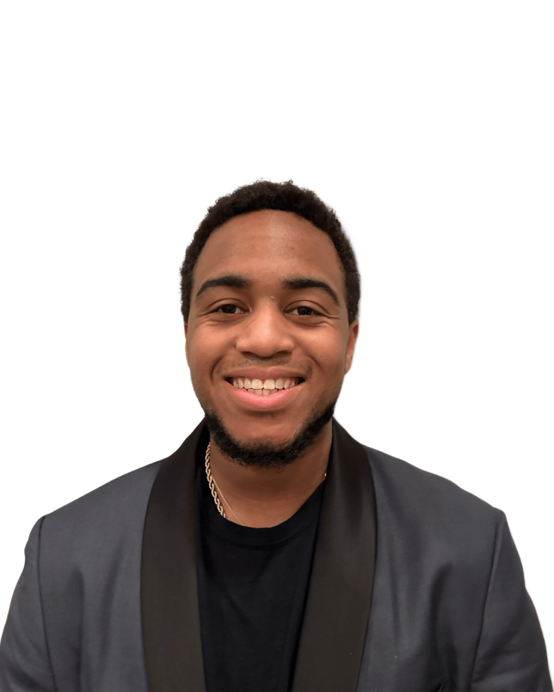 A person with a beard smiling while wearing a dark grey blazer over a black shirt against a white background.