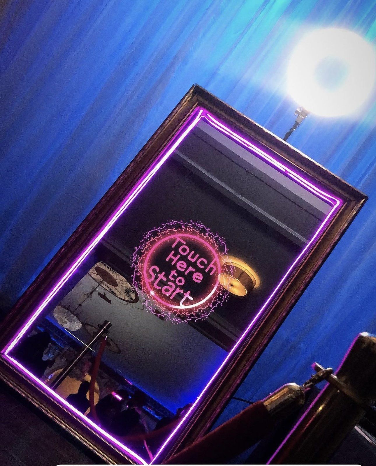 Neon-lit mirror photo booth in a blue room, reflecting a pink “Touch Me” sign.