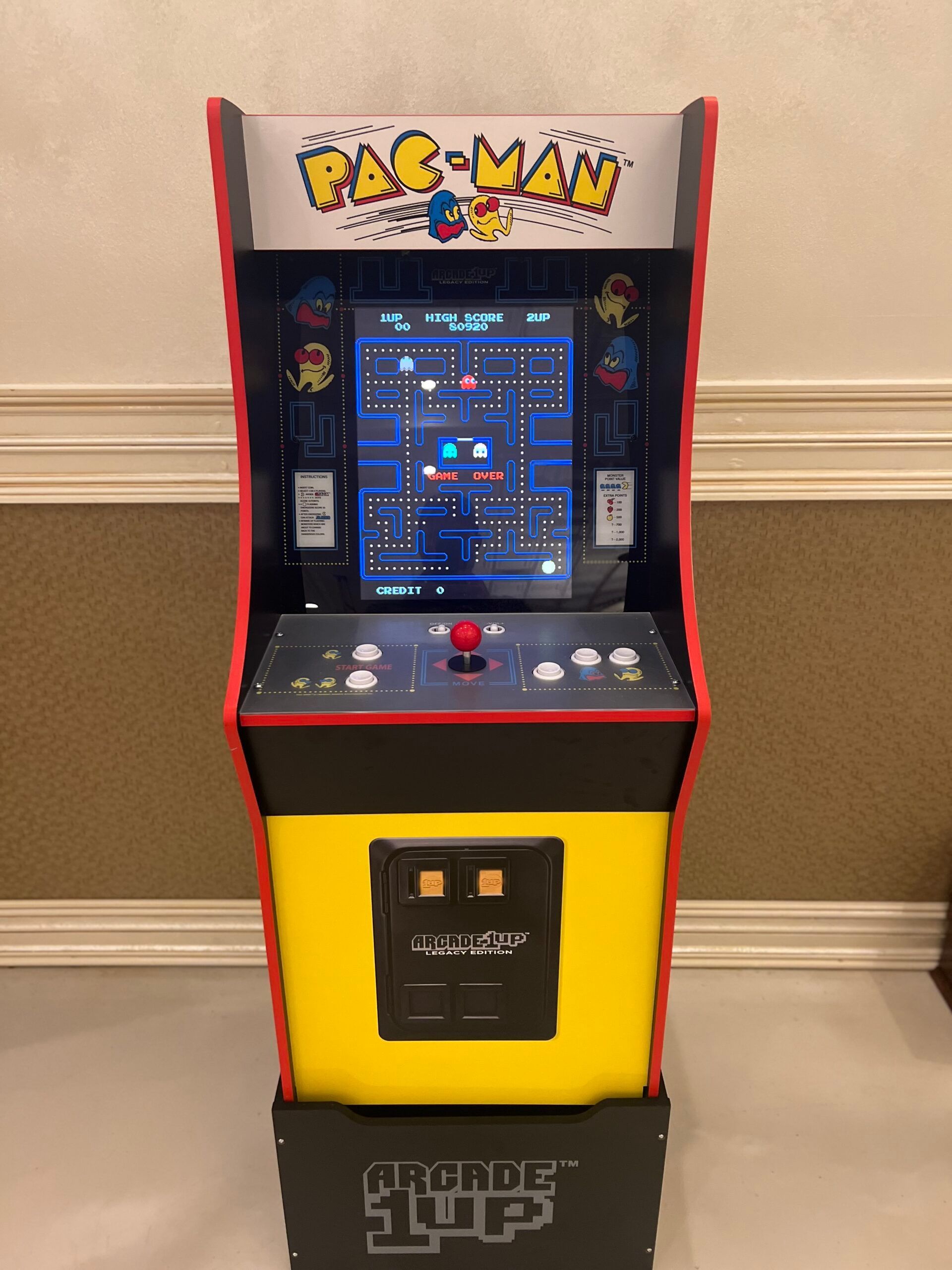 PAC-MAN arcade cabinet with red trim and a lit game screen in a room against a beige wall