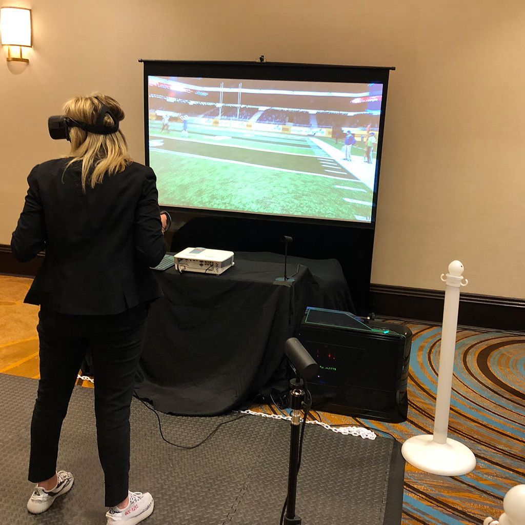 Guest playing a virtual reality football game using a headset and motion sensors with live gameplay on a projection screen.