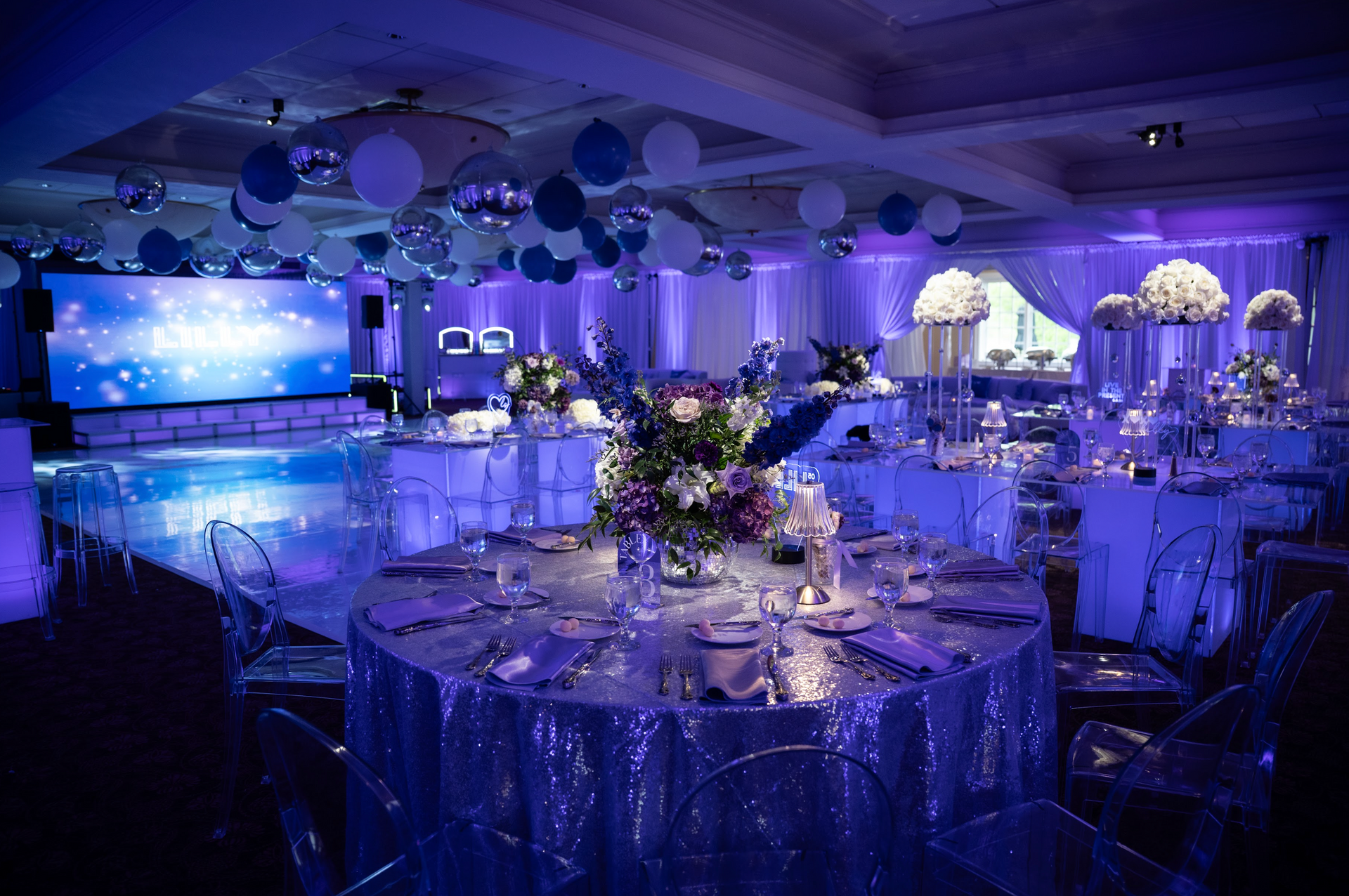 Purple-lit banquet hall with clear chairs, decorated tables, balloons, and a stage with a large screen.