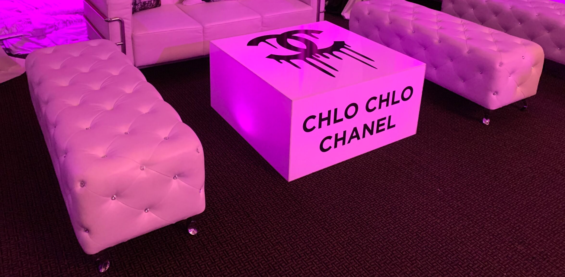 Custom Chanel-themed coffee table surrounded by white tufted benches and pink uplighting for a fashion-inspired lounge area.