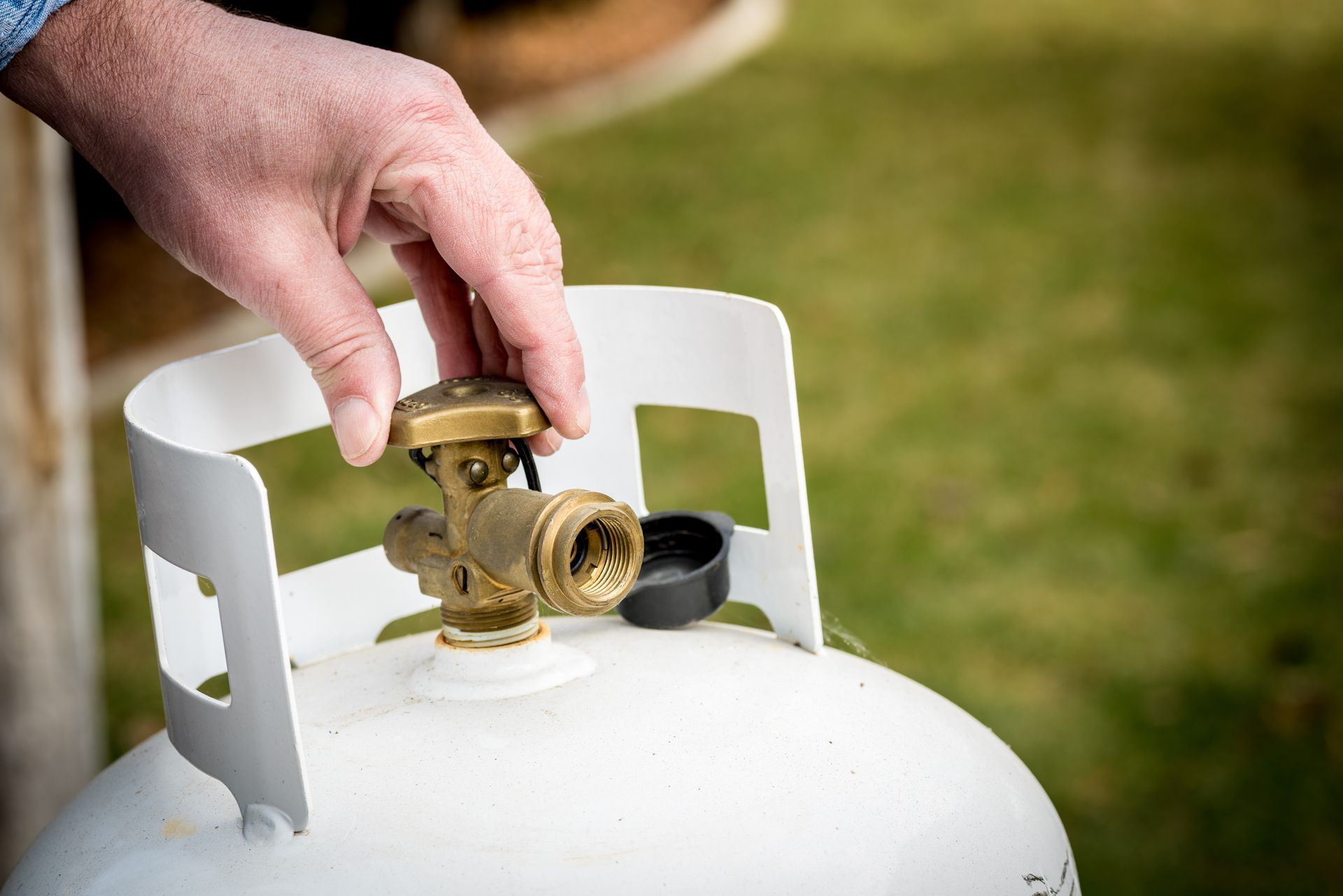 Hand adjusting the valve on a white propane tank set outdoors on green grass.