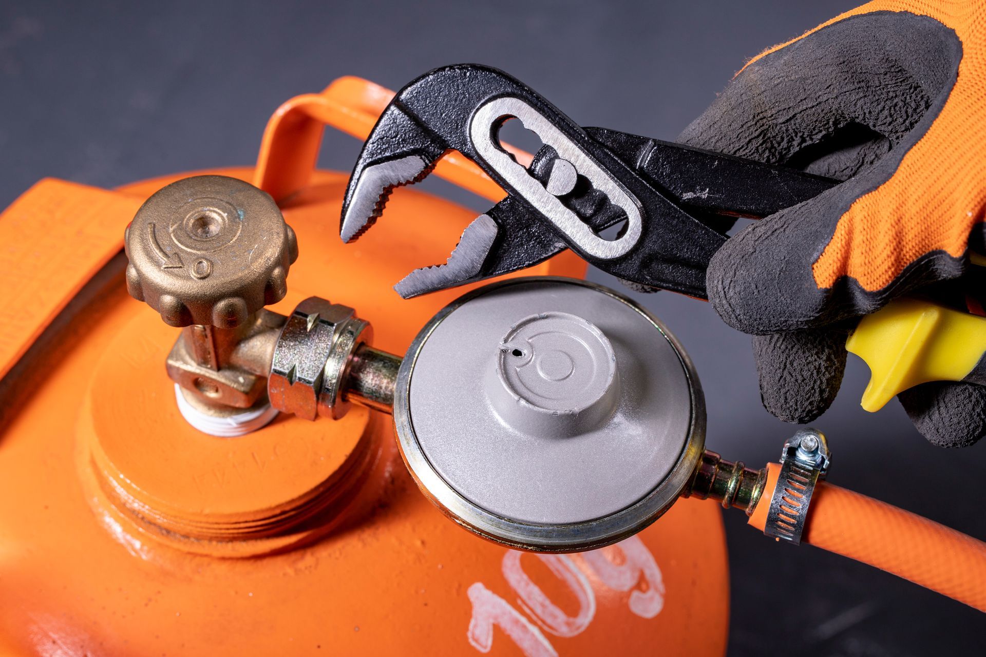 The gloved hands of a person holding a wrench above an orange gas tank.