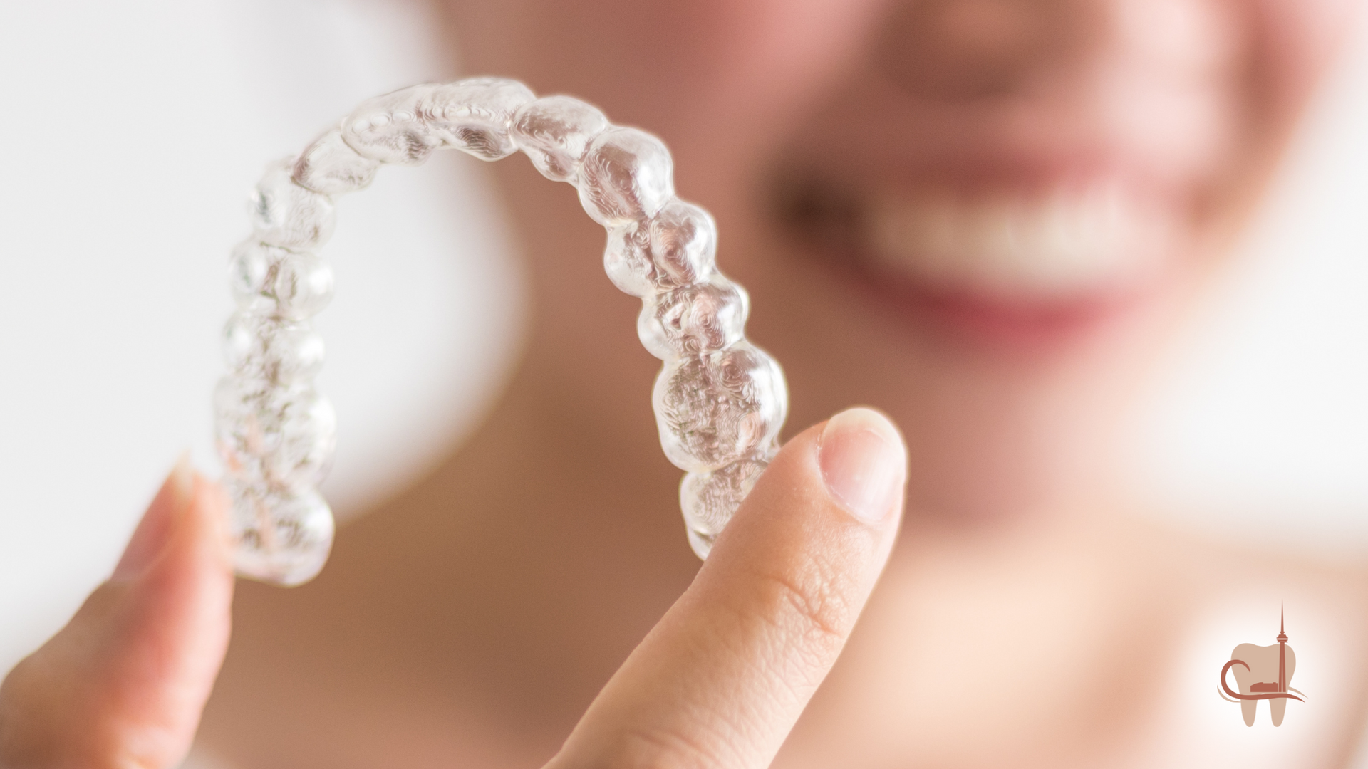 Person holding clear aligner; blurred smile in background.