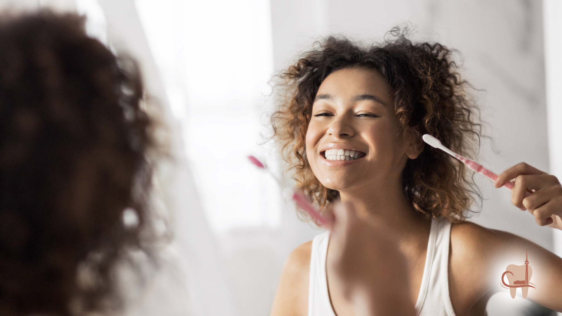 Woman brushing teeth in a bathroom, smiling at her reflection. White tank top, curly brown hair, pink toothbrush.