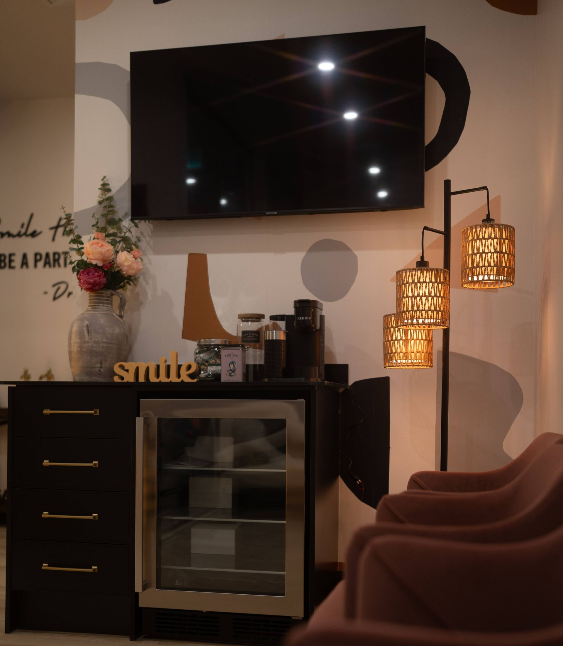A waiting area with a TV, coffee bar, pink chairs, and a lamp. Neutral color palette,