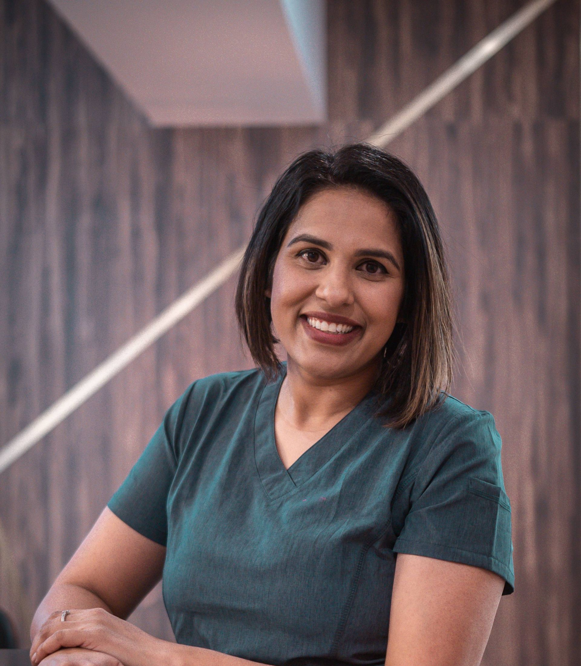Woman with dark hair smiling, wearing teal scrubs, set against a wood-paneled wall.