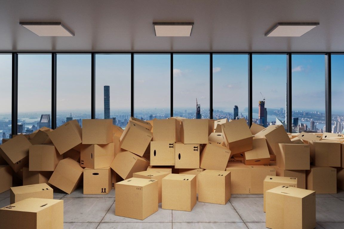 Cardboard boxes stacked in a bright office with floor-to-ceiling windows and a city skyline beyond.