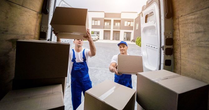 Two movers in blue overalls carry boxes into a truck from a warehouse doorway.