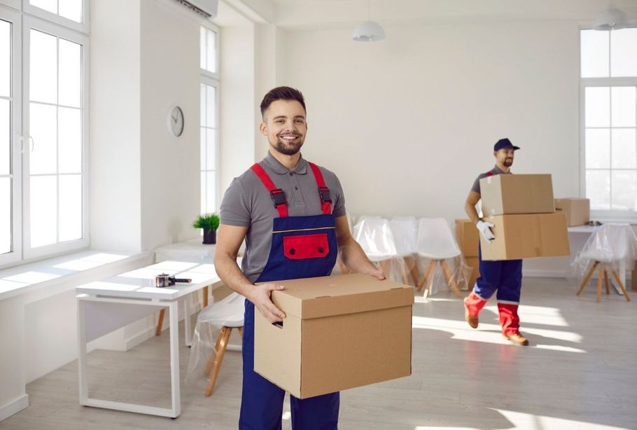 Two movers carrying cardboard boxes in a bright, white room with large windows