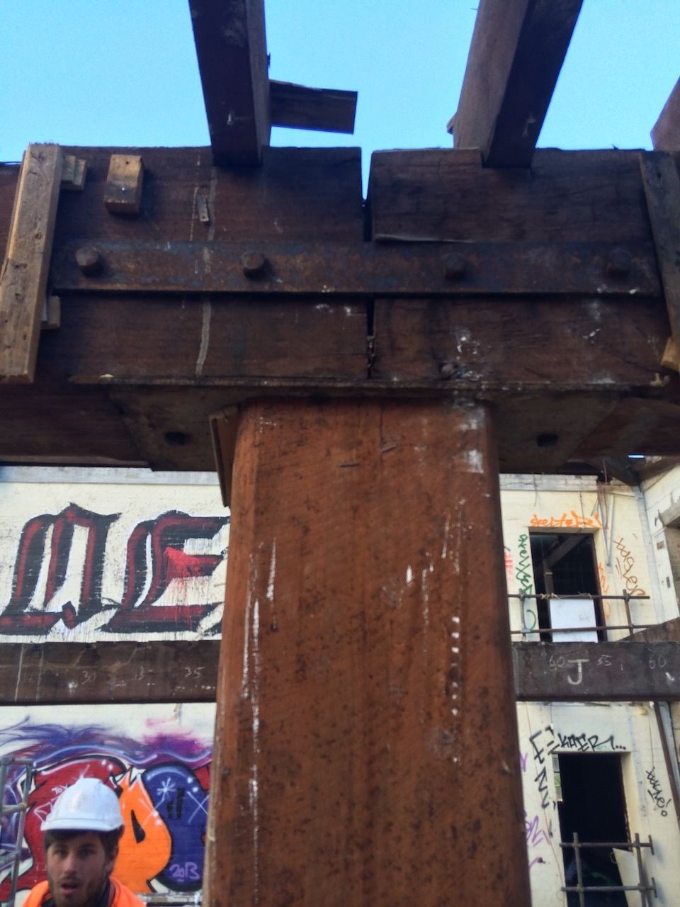 Construction site: a metal beam attached to a square metal column. Worker in hard hat, orange vest at bottom in Brisbane.