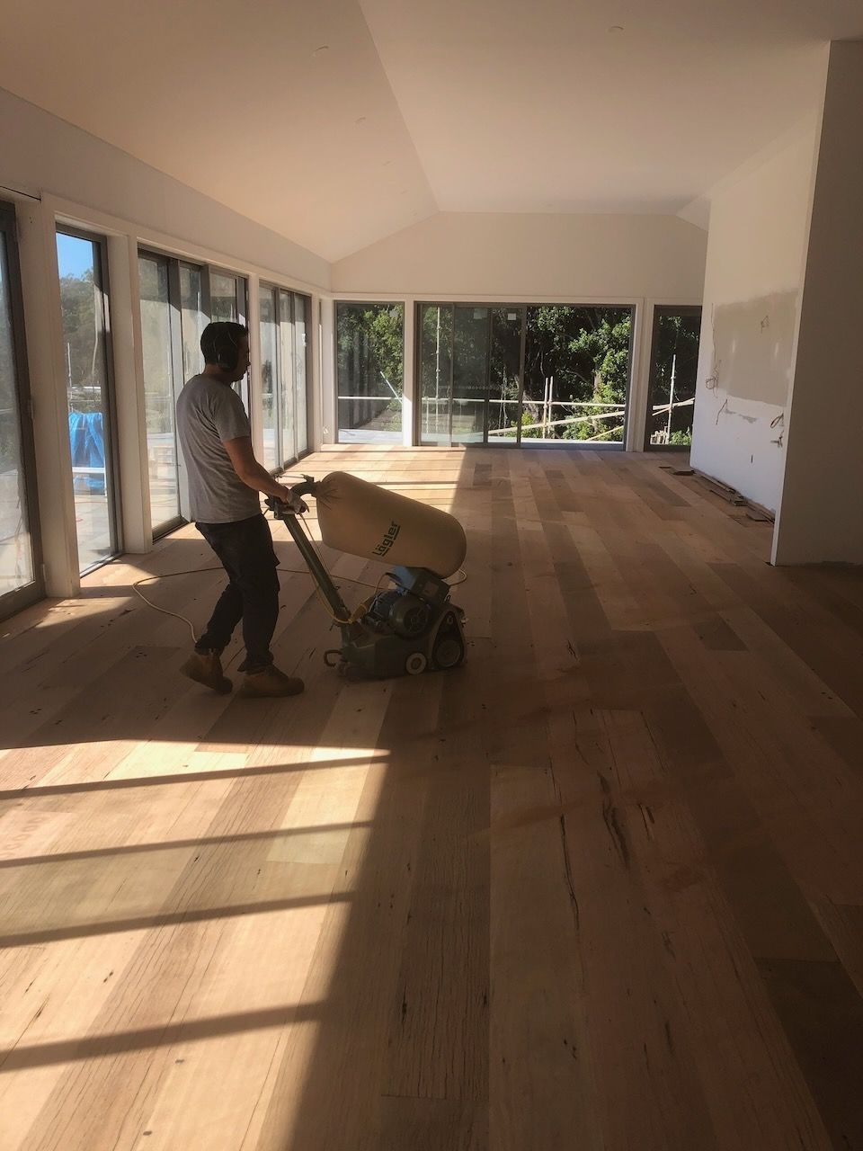 Man sanding a wooden floor in a sunlit room with large windows in Brisbane.