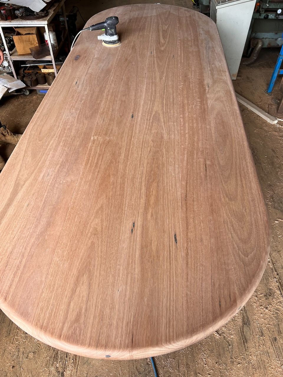 Oval wooden table being sanded with a power sander; sawdust visible.