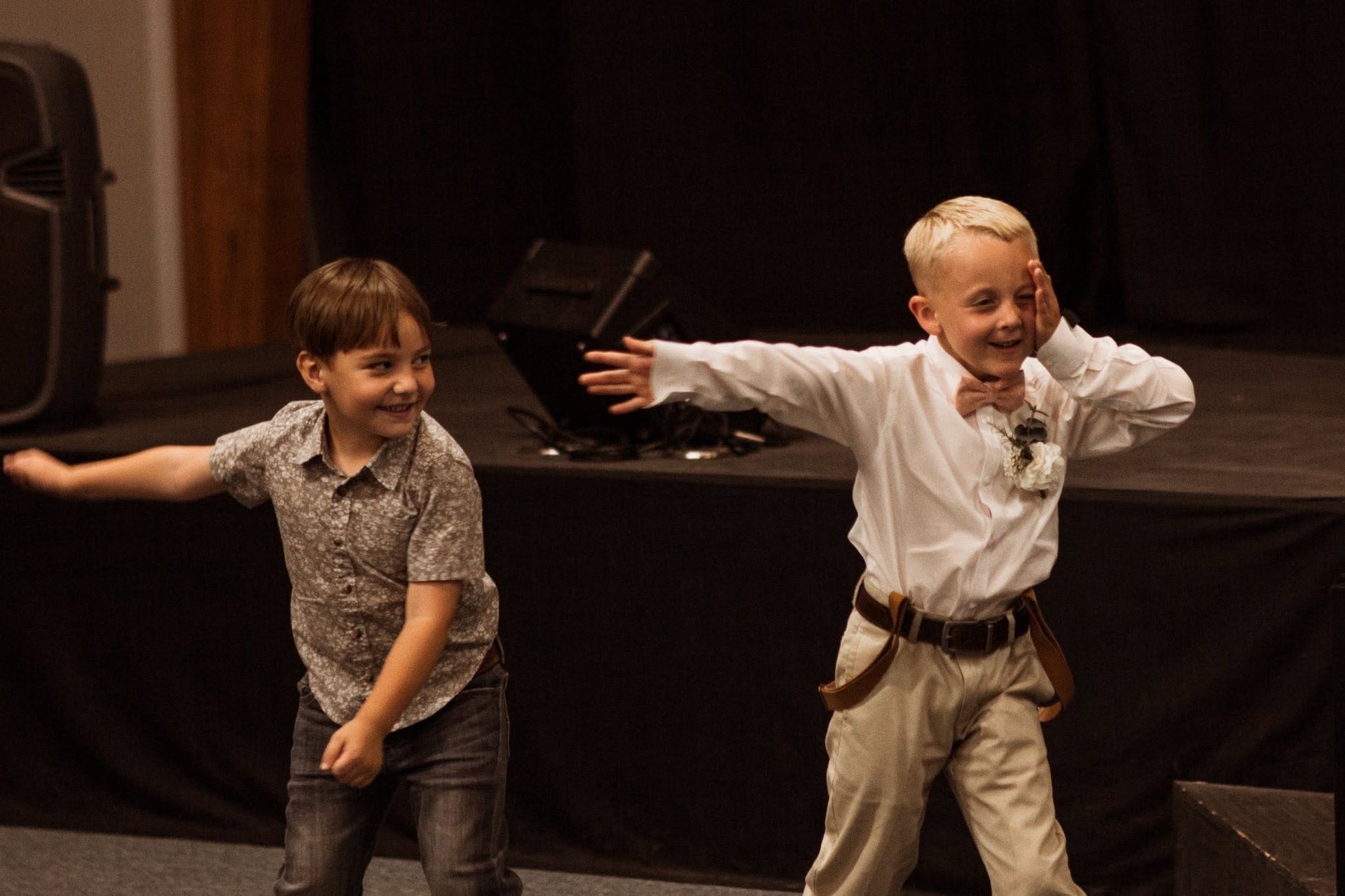 Two children dancing on a dim stage, arms outstretched and smiling