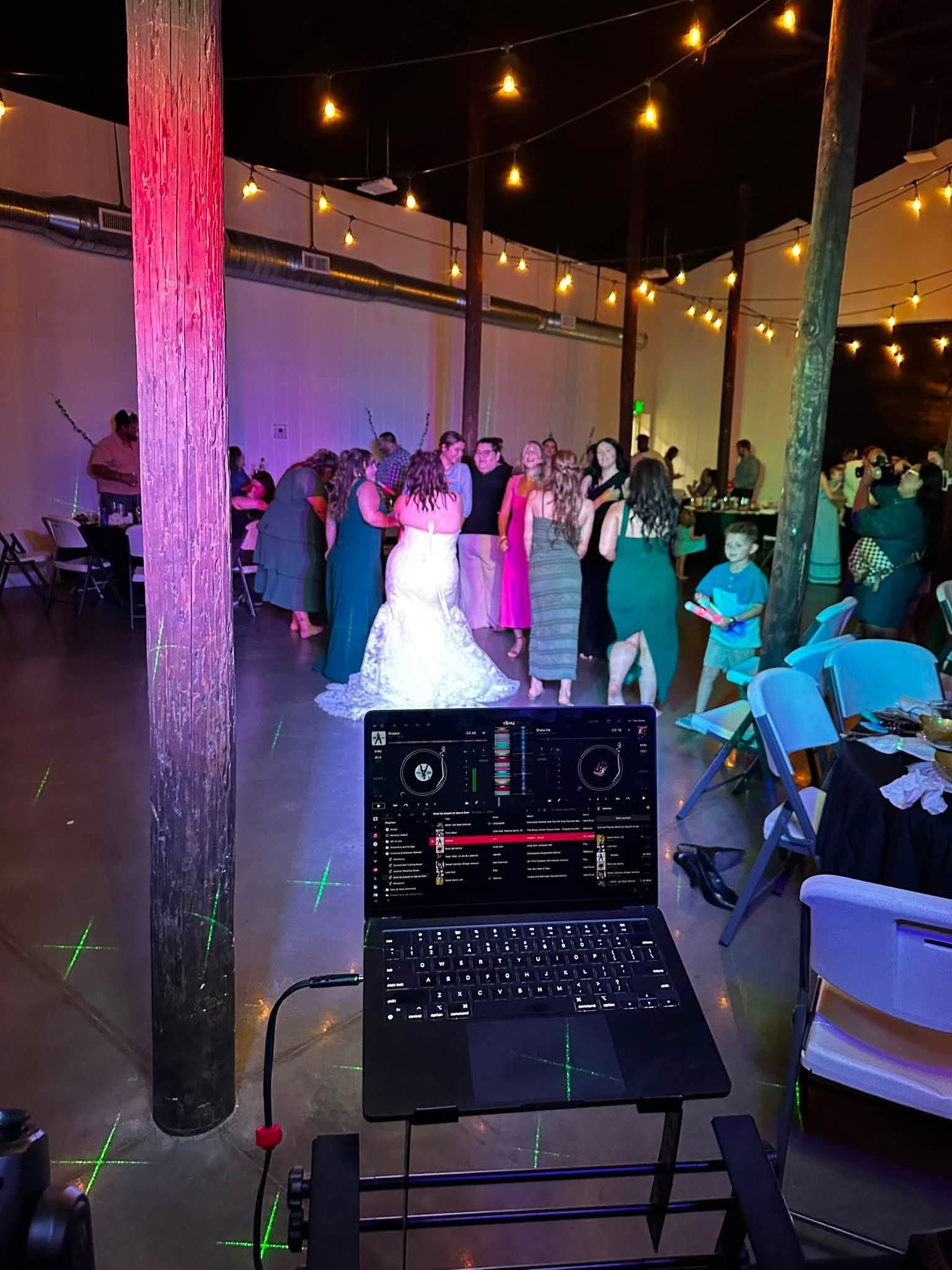 Outdoor wedding reception with guests dancing under string lights, viewed behind a DJ laptop and speaker.