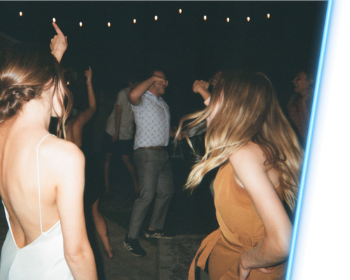 People dancing at a dimly lit party, with two women in the foreground and string lights overhead.