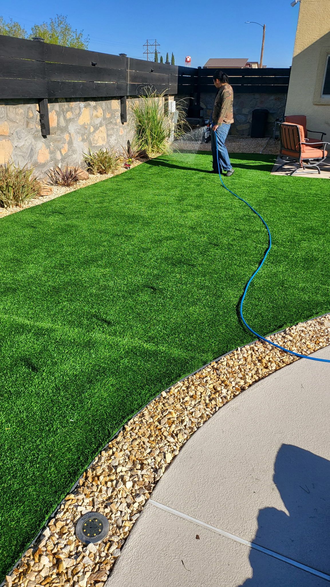 Backyard with bright green lawn, stone patio, and a person watering plants near the fence