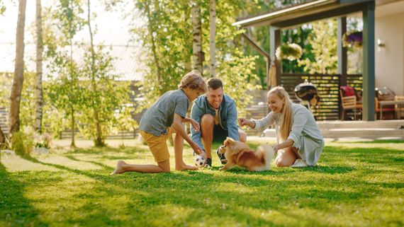 Two adults and two children petting a small dog on a sunlit lawn beside a house