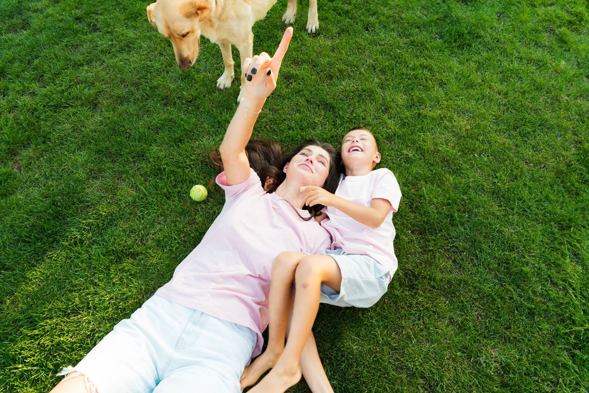 Two people lying on grass, smiling and reaching up to pet a dog nearby